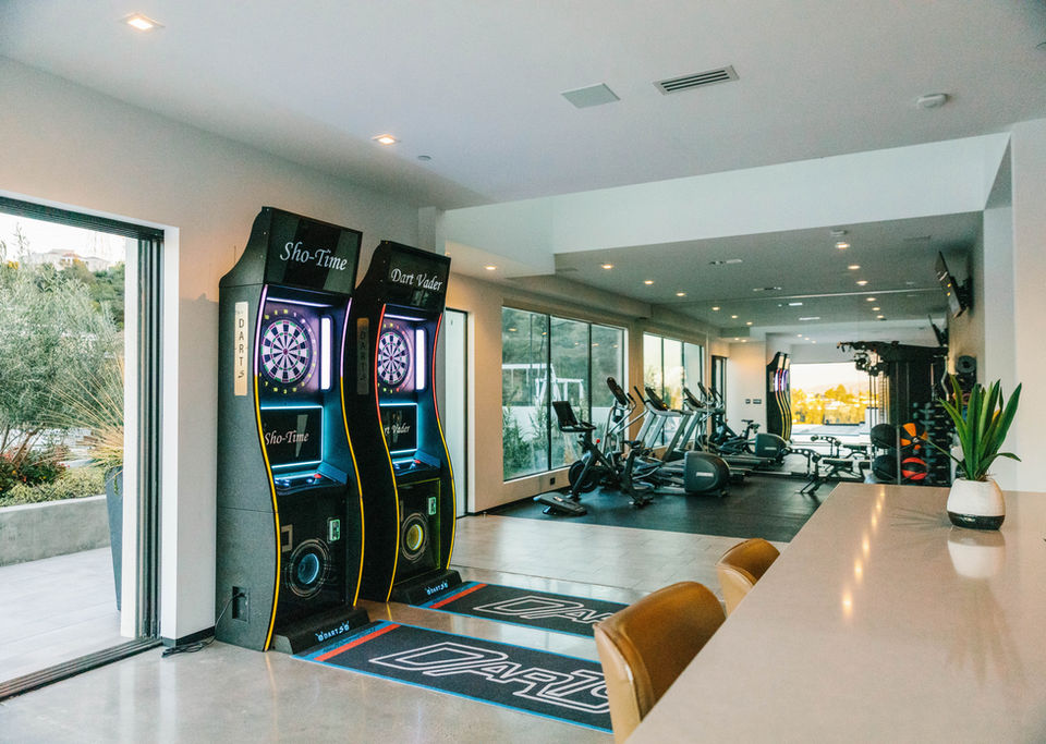 A modern entertainment and fitness suite in a Los Angeles County home featuring electronic dartboards and a variety of gym equipment, designed by Leli Construction.