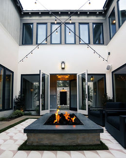 A contemporary courtyard in Costa Mesa featuring a central stone fire feature, large glass doors, string lights, and elegant tile flooring, designed by Leli Construction.