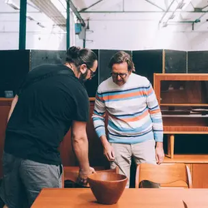 Twee bezoekers bekijken een houten schaal tussen teakhouten dressoirs en vitrinekasten in een industriële hal