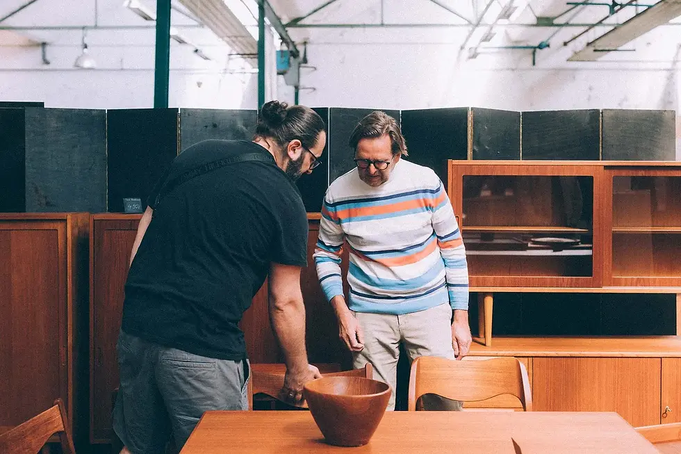 Twee bezoekers bekijken een houten schaal tussen teakhouten dressoirs en vitrinekasten in een industriële hal