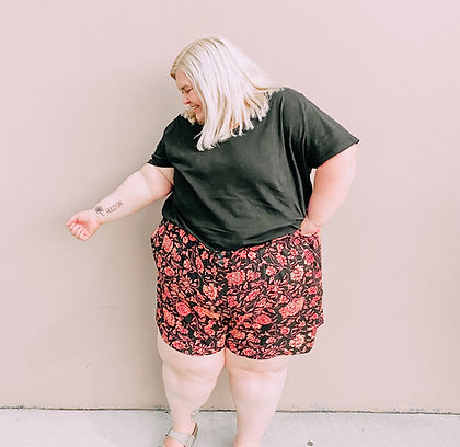 A woman stands in front of a tan wall in black and orange looking at a tattoo on her forearm