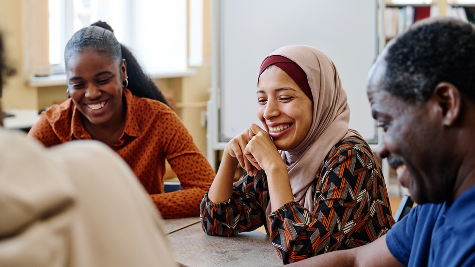 Smiling students in a classroom sit around a table, chatting and laughing in a warm, bright group discussion