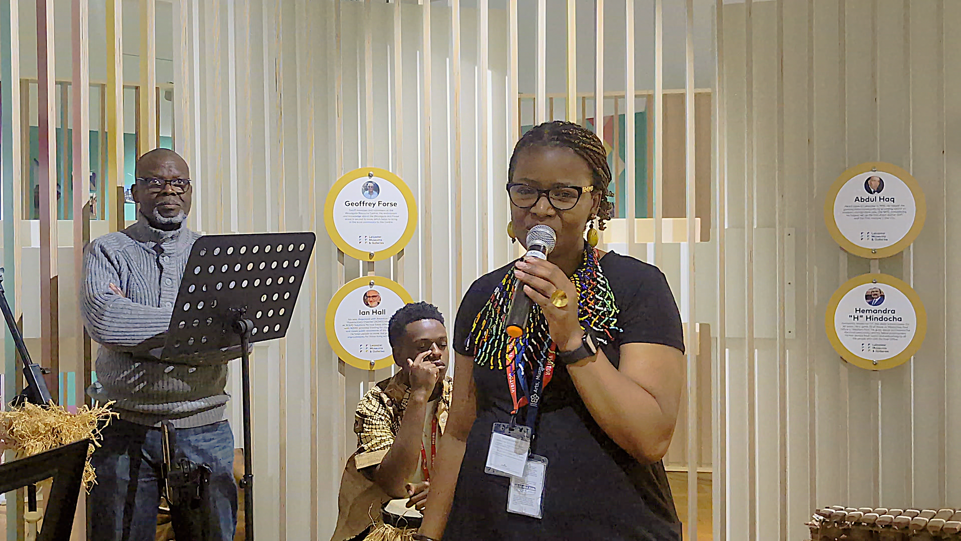 A woman sings into a microphone in front of a small audience. Behind her, cultural heritage profiles are displayed on circular panels attached to wooden slats.