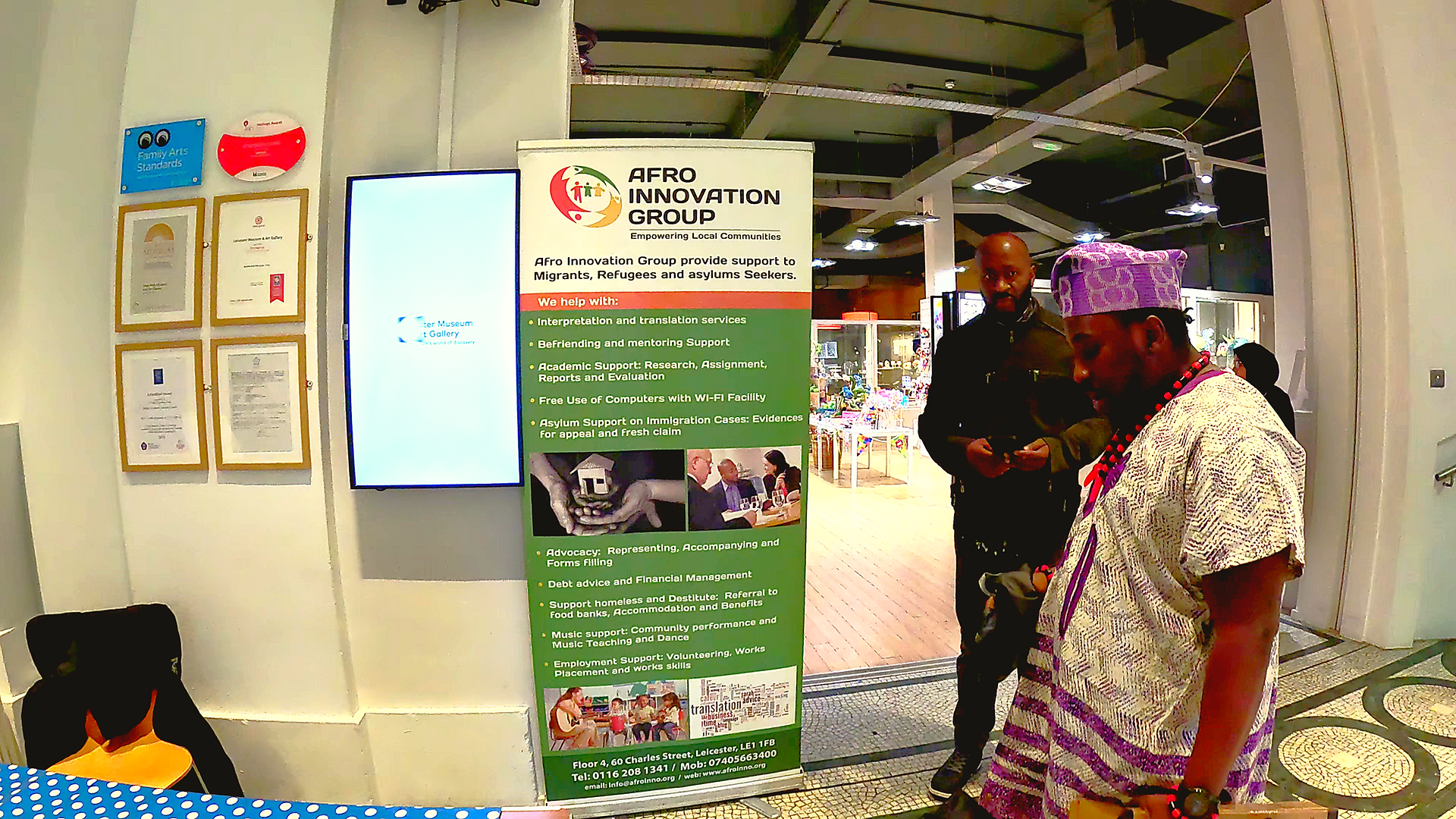A man in traditional African attire stands beside an Afro Innovation Group banner and talks to another visitor. The museum’s certification plaques and digital welcome screen are visible in the background.