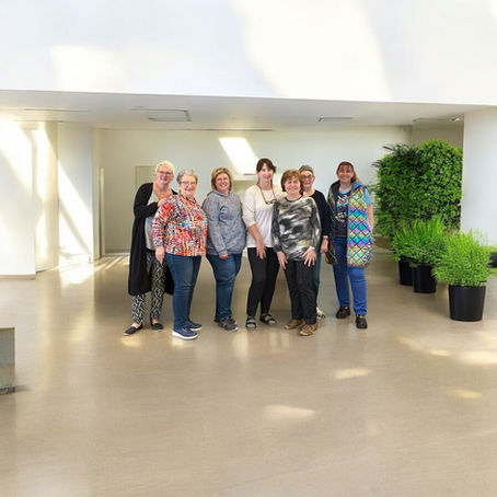 Sieben Frauen stehen nebeneinander und lächeln in die Kamera. Gruppenfoto einer Nähwoche in einem hellen Raum mit Pflanzen im Hintergrund. 