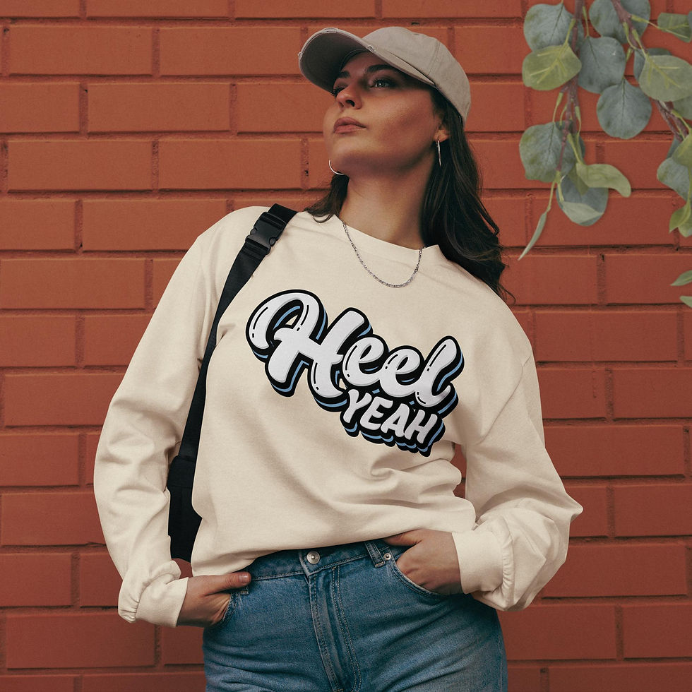 Woman flaunting a cream-colored (ecru), relaxed, heavyweight, long-sleeve shirt, showcasing a Carolina-themed, Heel Yeah Design in front of a brick wall.