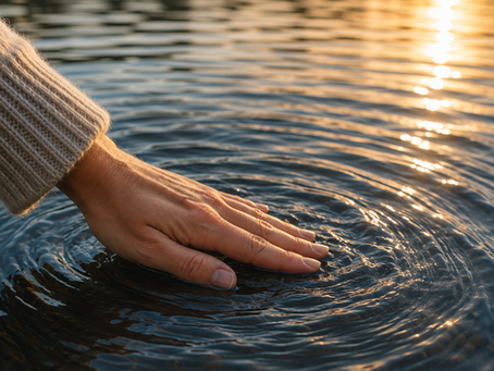 Hand in a knit sleeve touches tries to smooth the rippling water at sunset, with golden reflections on a rippling lake.
