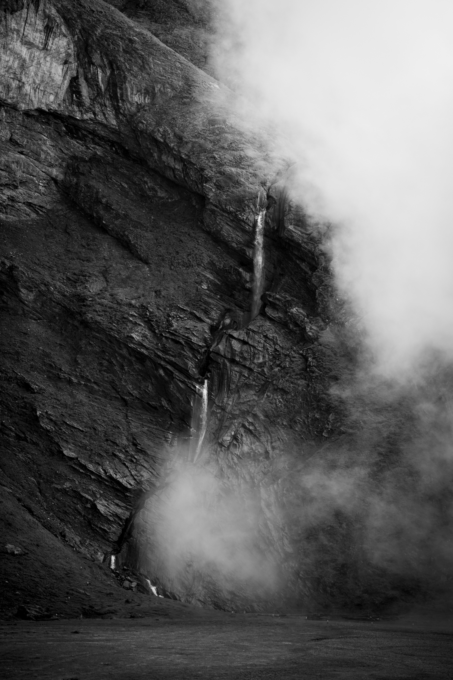 Ein schmaler Wasserfall zieht durch dunkle Felswände. Nebel verhüllt den unteren Teil und löst die Landschaft in Grautöne auf.