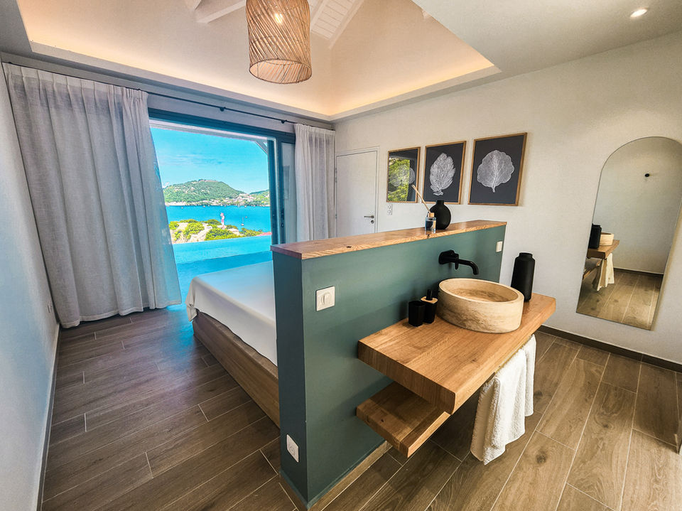 Chambre à coucher avec une fenêtre ouverte donnant vue imprenable sur la mer et une piscine extérieure. La pièce est élégamment meublée avec un grand lit et des détails modernes. À l'arrière de la chambre, une salle de bain lumineuse, équipée d'une douche et de commodités modernes. Les couleurs de la chambre sont chaudes (bois).