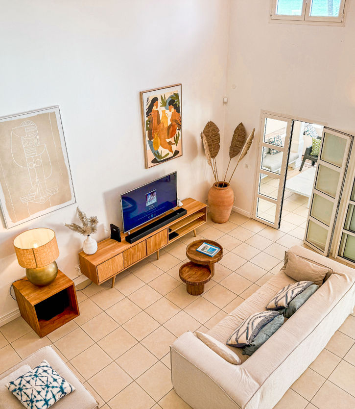Vue aérienne du salon spacieux et lumineux d'une villa Caraïbe Location, avec un mobilier moderne, un canapé, une télévision, des éléments décoratifs et des baies vitrées offrant une vue panoramique.