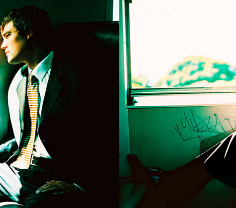 Homem de casaco azul sentado dentro de um comboio, com a cabeça baixa e uma mochila ao lado, representando a frustração e a espera.