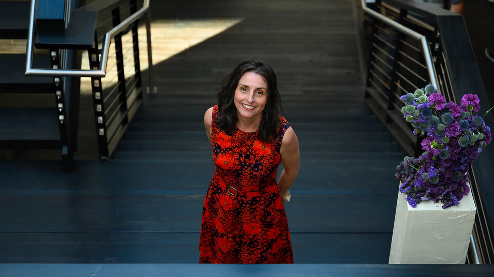 Woman in a red floral dress smiles while standing on a dimly lit staircase. A bouquet of purple flowers is visible on the side.