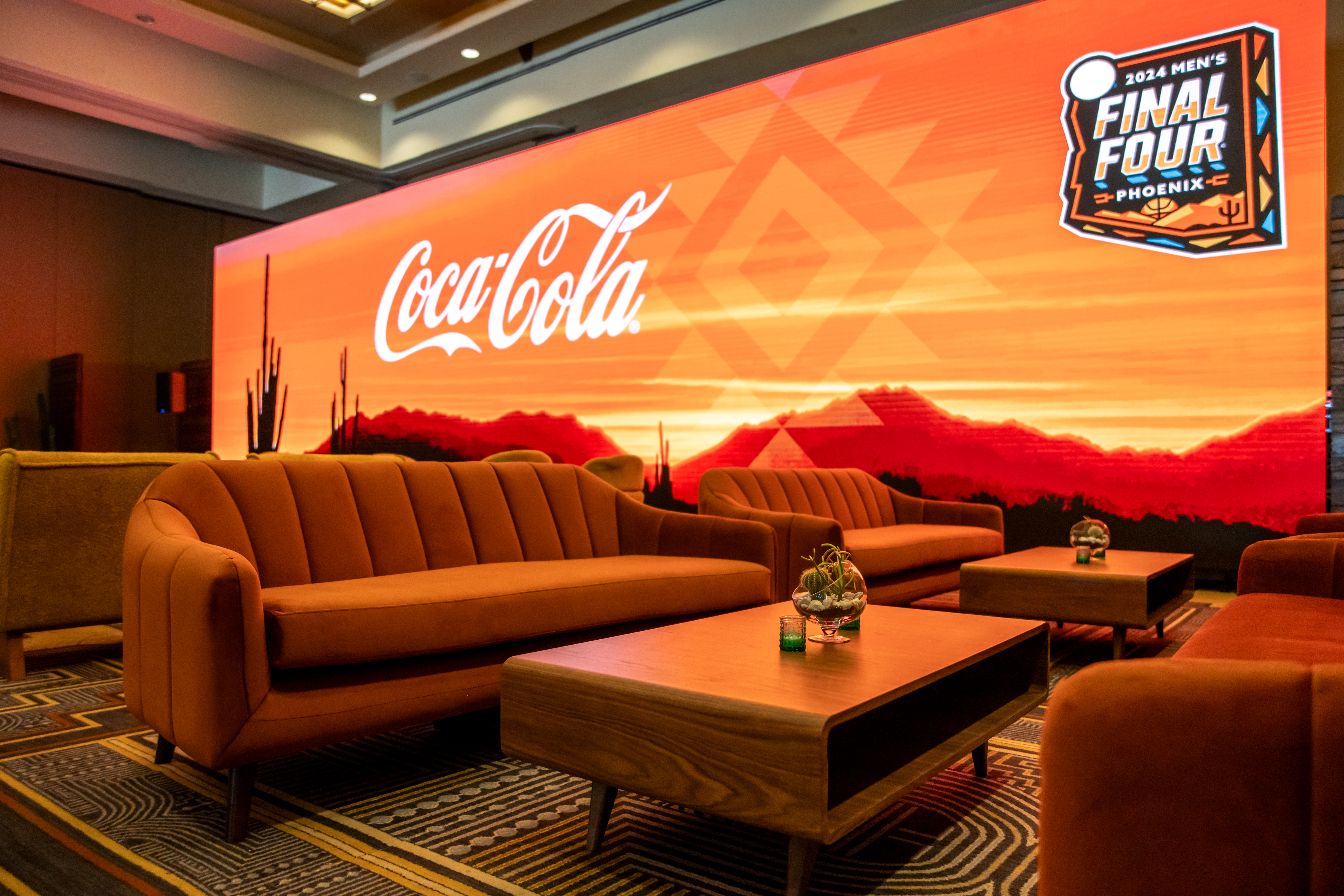Coca-Cola guest lounge with Southwest-themed furniture and LED video wall displaying 2024 Final Four logo.
