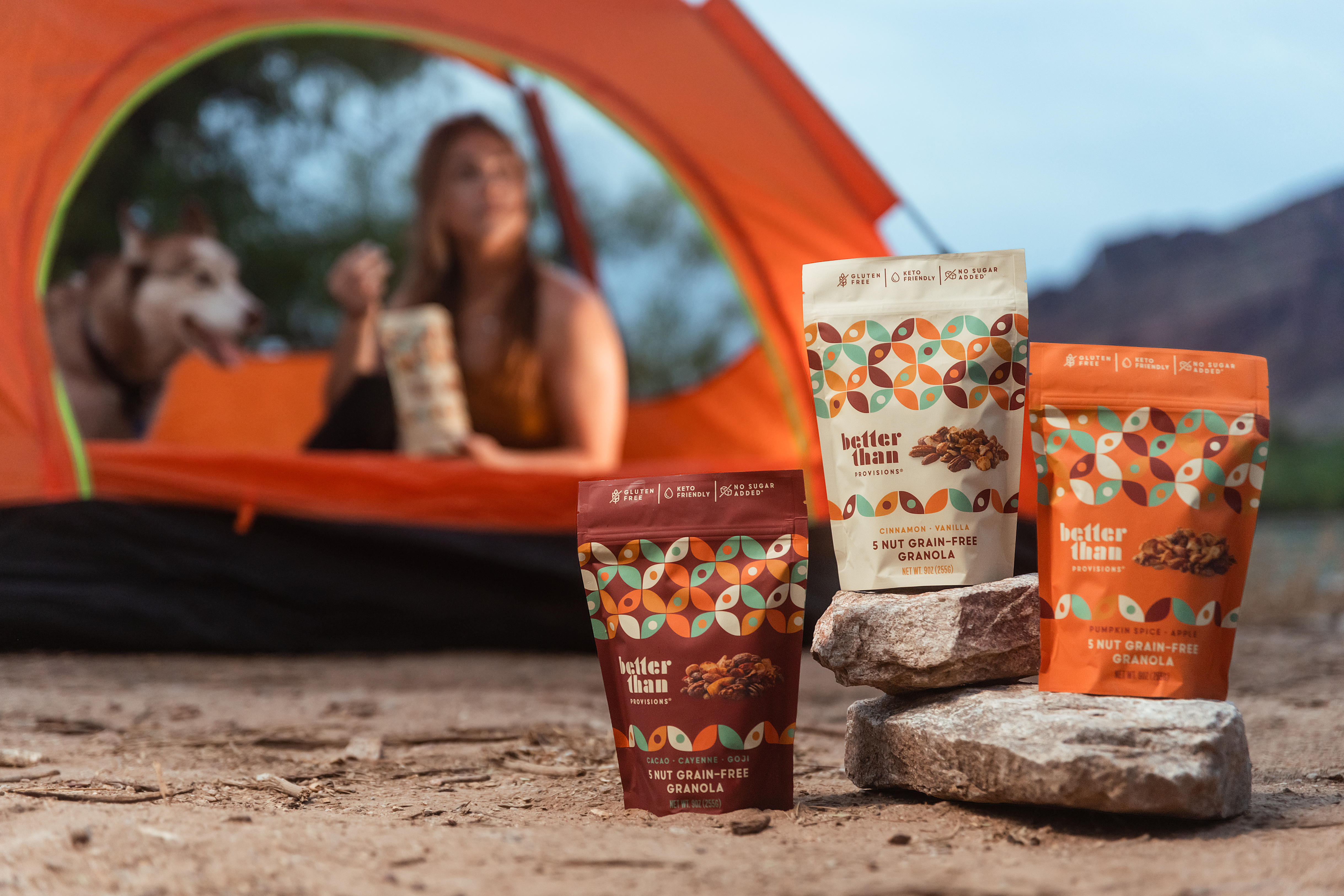 Line of three Better Than Provisions granola flavors with tent and person with dog in background, promoting active lifestyle.
