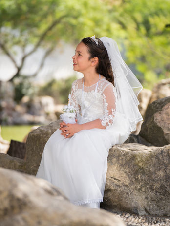 Girl wearing a white communion gown and veil sitting on rocks during an outdoor photo session.