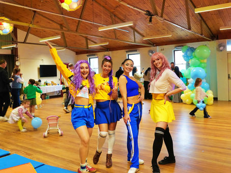 Four smiling women in colorful K Pop Demon Hunter outfits pose at a kids' party in a hall, with balloons and children playing behind them.
