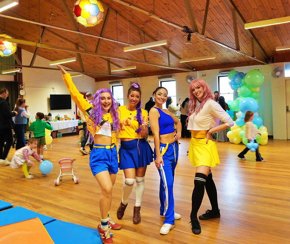 Four smiling women in colorful K Pop Demon Hunter outfits pose at a kids' party in a hall, with balloons and children playing behind them.