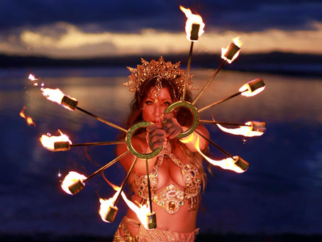 A woman in sparkling attire performs fire dancing by a lake at sunset. She holds two fire fans, creating an intense, dramatic scene.
