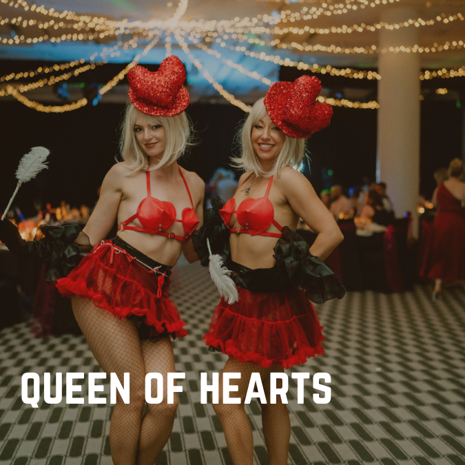 Two women in red heart costumes and hats pose smiling, holding feathers. Party setting with string lights; "QUEEN OF HEARTS" text visible.