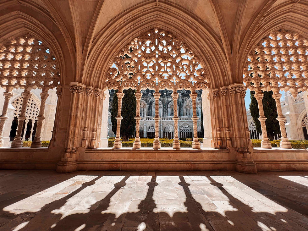 Ornate arches with intricate patterns frame a historic building and trees outside. Sunlight casts detailed shadows on the stone floor.