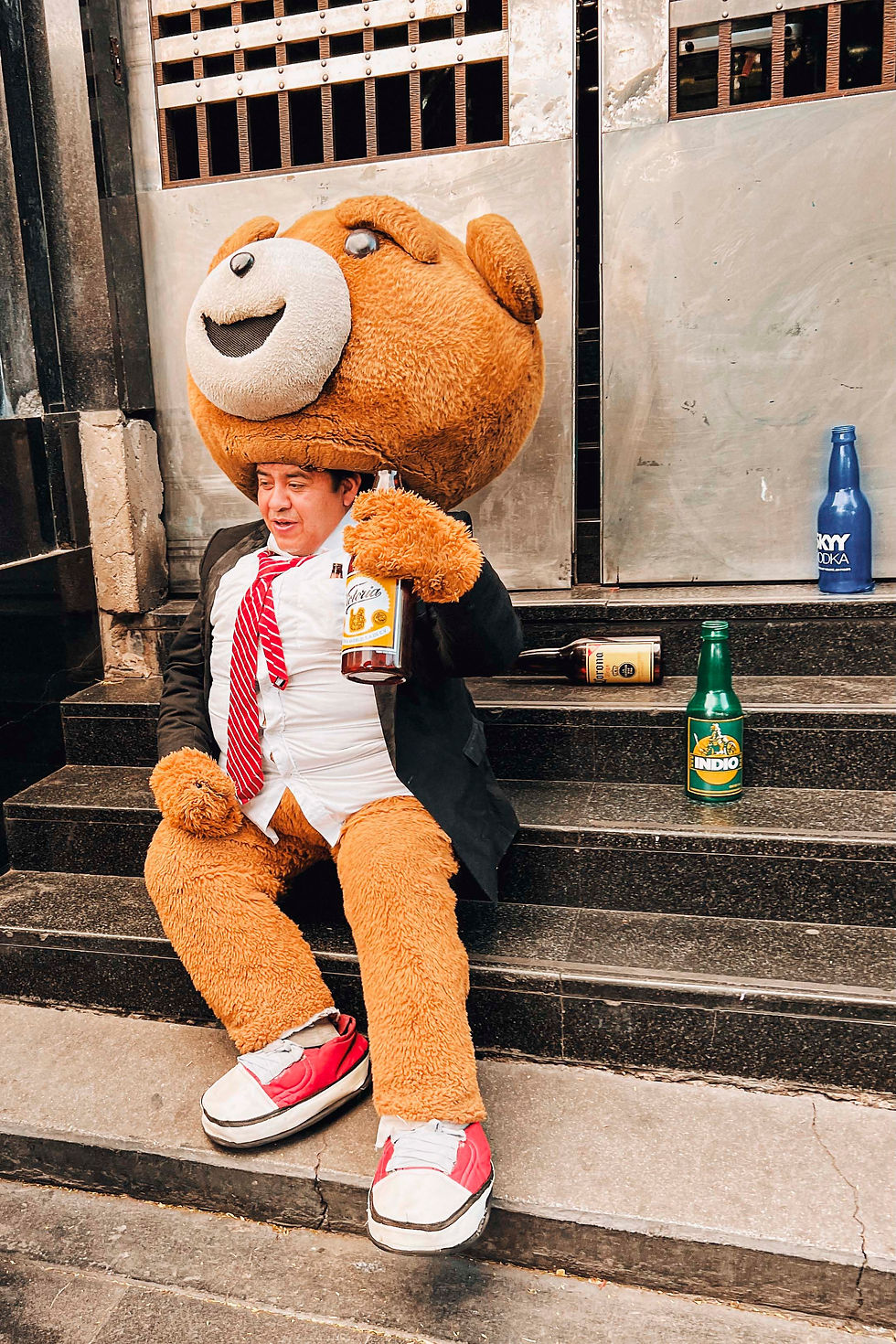 Man in bear costume sits on steps, holding a beer bottle. Dressed in a suit with a red tie. Vodka and beer bottles nearby. Urban setting.