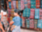 Women in traditional clothing stand near vibrant, patterned textiles at an outdoor market. One holds a black bag. The mood is busy and colorful.