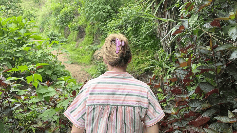A person with a striped shirt and purple hair clip walks on a lush green forest path. The mood is peaceful and natural.