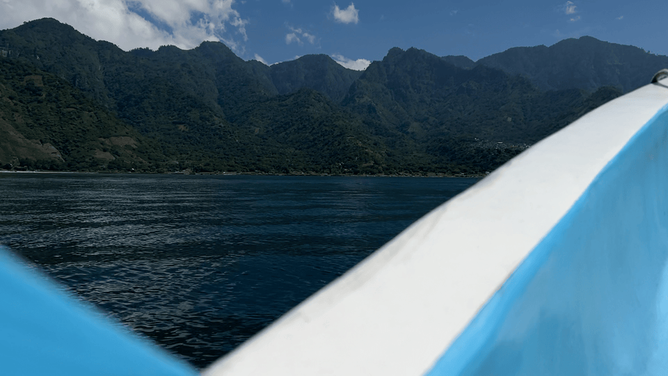 Boat on blue water with lush green mountains in the background under a partly cloudy sky. The boat railing is white and blue.