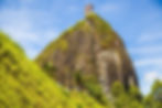 Steep stairway climbs a massive rock in Guatapé, Colombia, where lush greenery surrounds. Bright, sunny day under a clear blue sky. A few people seen ascending.