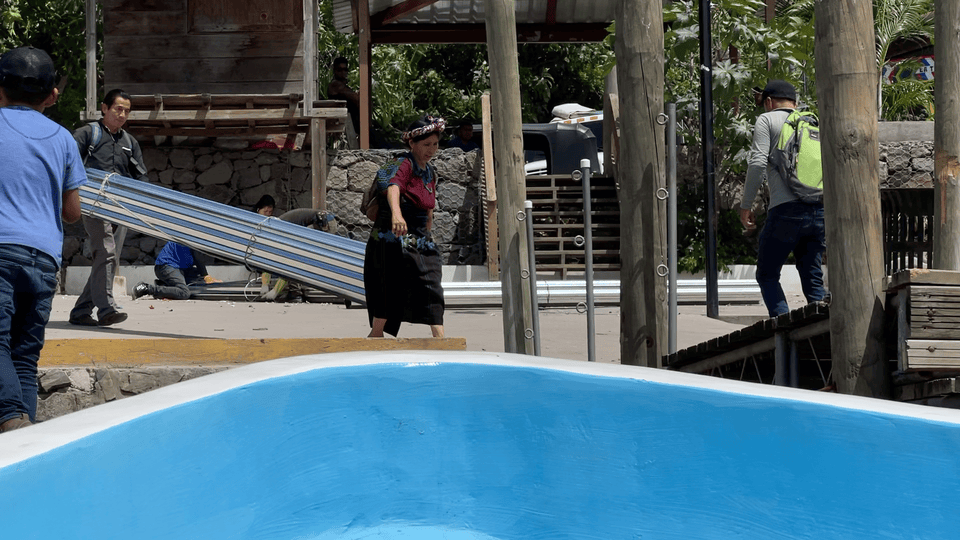 Men carry metal sheets and a woman walks near a blue-bordered pool. Wooden structures and greenery in the background, sunny day.