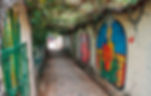 Narrow alley with colorful murals on walls, featuring a seated figure. Overhead vines create an arch; a green gate is on the left.