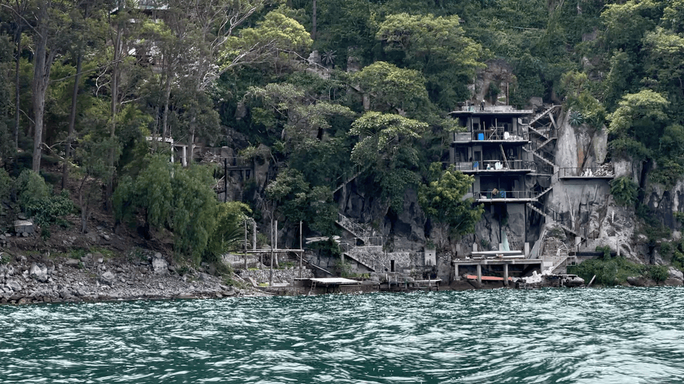 Multi-story house built into a lush, rocky hillside by a lake. Trees surround the structure, and boats are docked at the shoreline.