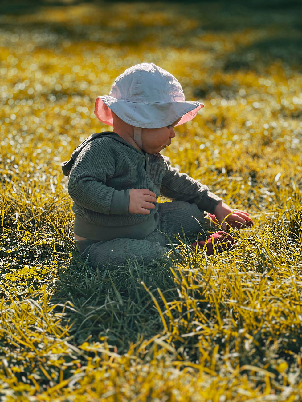 Baby in a white hat sits on green grass, touching the ground. Sunlight creates a warm, serene atmosphere in the outdoor setting.