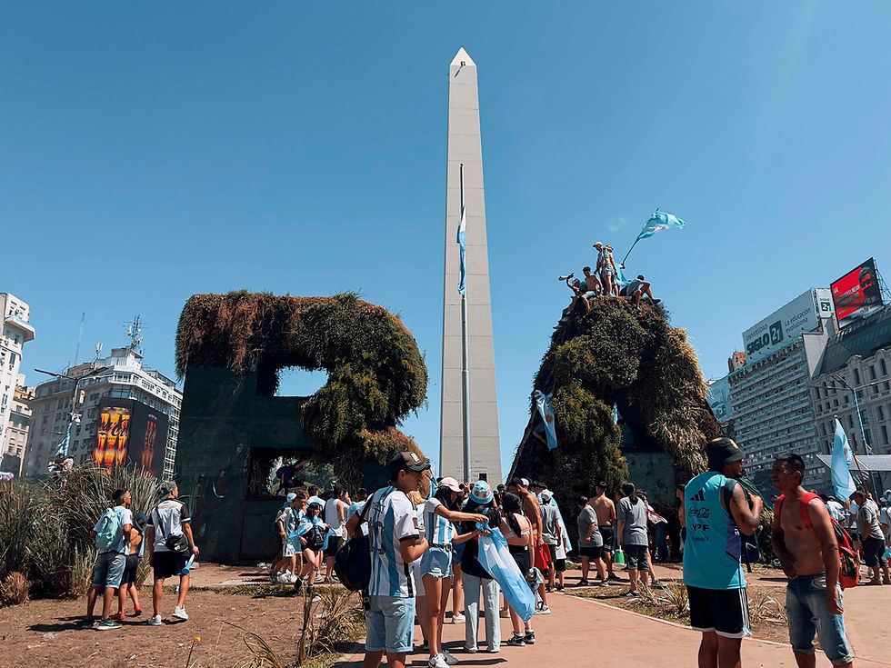 People gather around the Obelisk in Buenos Aires, some climbing a grassy structure, waving blue flags. The scene is sunny and lively.