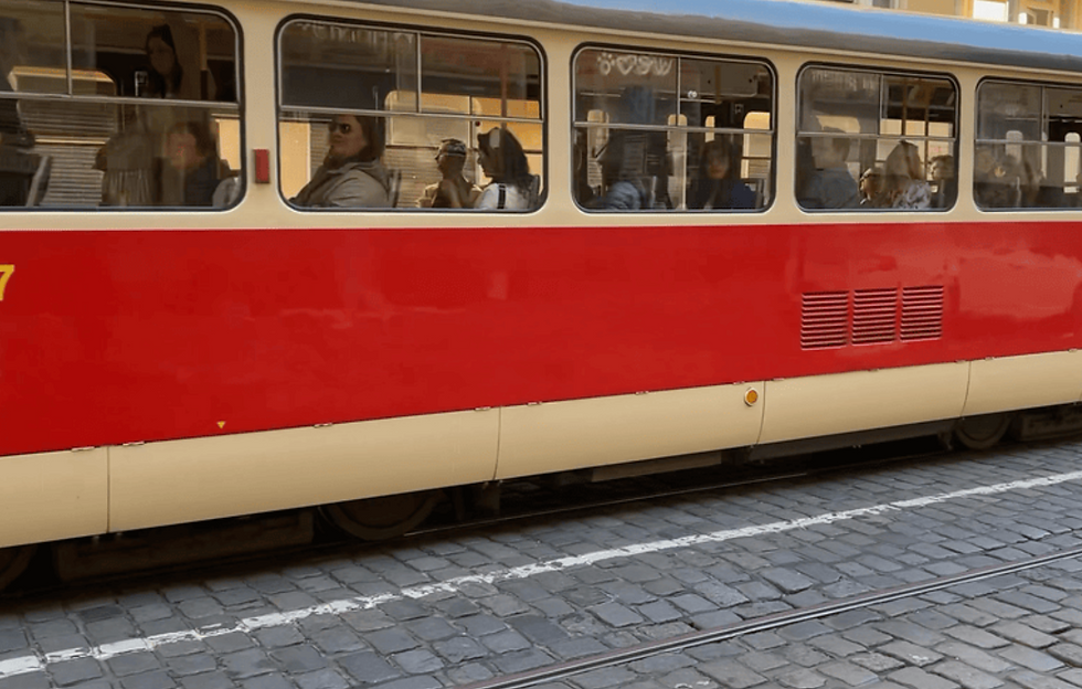 Red tram with passengers travels on cobblestone street. People appear relaxed. Large windows show an urban setting in daylight.