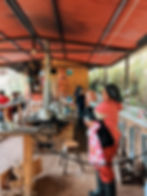 Woman in pink holds baby in a rustic kitchen with red roof. Pots on stove, two people in background. Cozy, homely atmosphere.