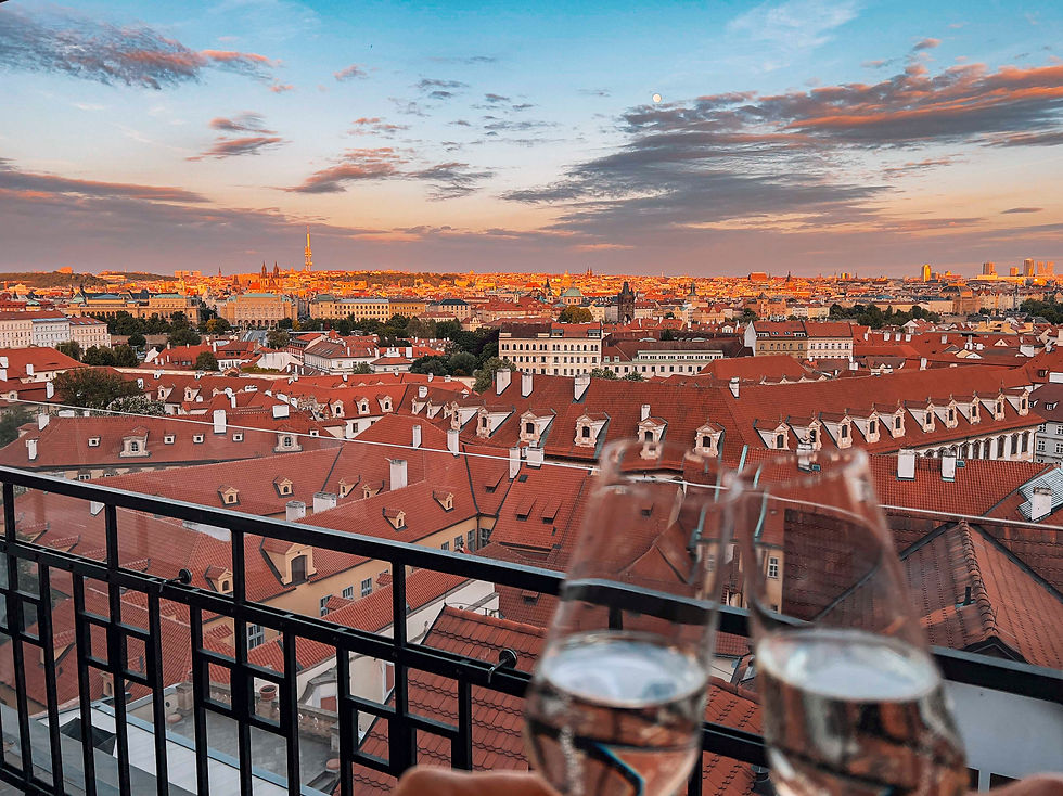Two champagne glasses clink on a balcony overlooking a cityscape with red roofs at sunset, under a partly cloudy blue sky.