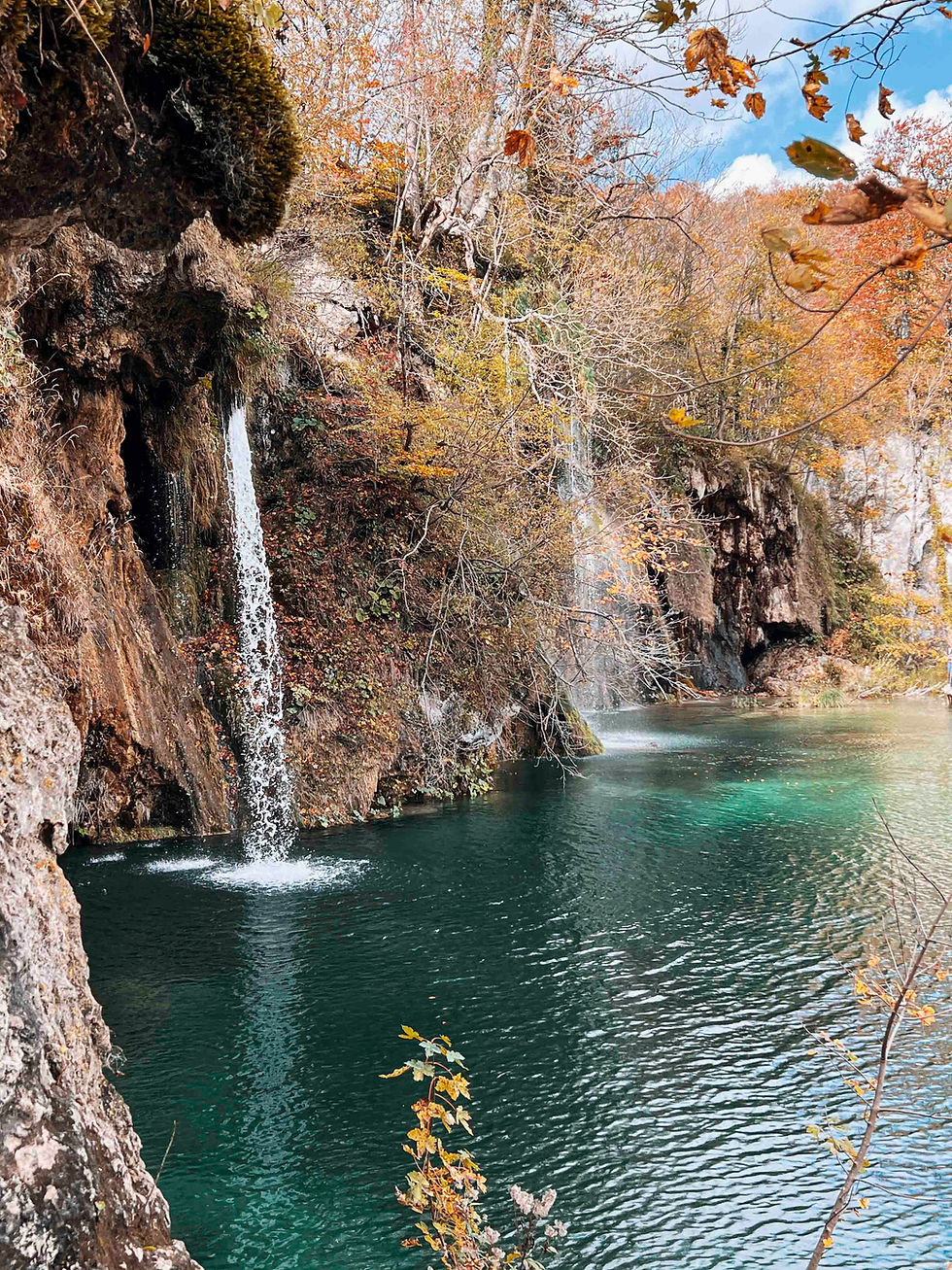 Waterfall cascading into a clear turquoise pool, surrounded by autumn trees with orange leaves, creating a serene and picturesque nature scene.