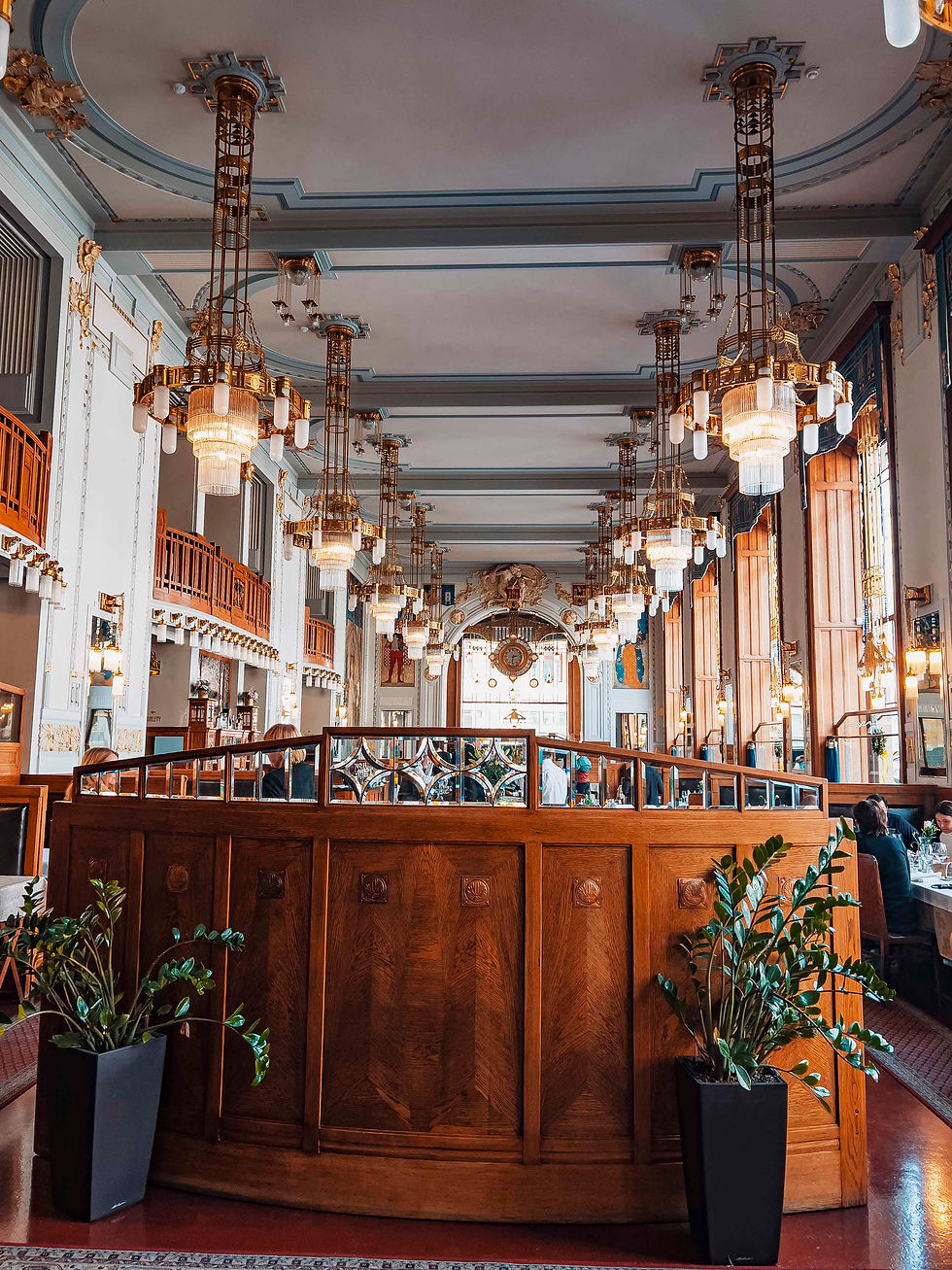 Elegant restaurant with ornate chandeliers, wooden decor, and lush plants. Diners sit at tables along the sides, creating a historic ambiance.