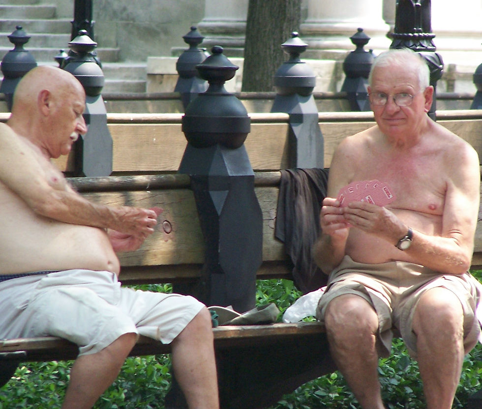 Two shirtless elderly men play cards on a park bench. One looks focused, the other glances up. Surrounded by greenery and lampposts.