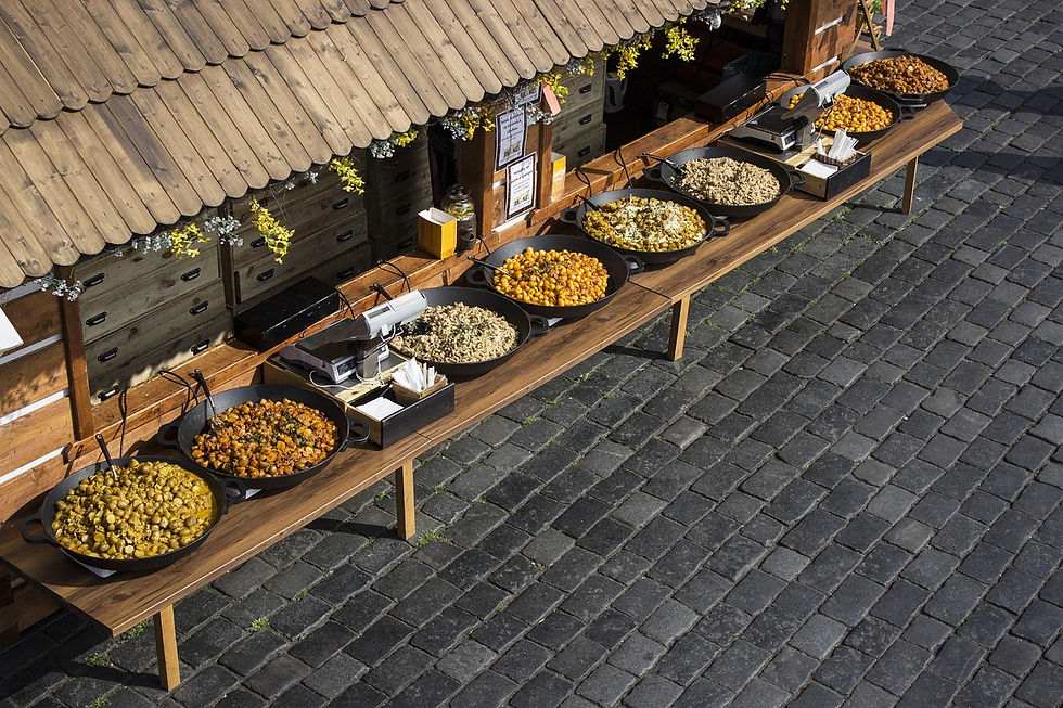 Large pans of colorful food displayed on a wooden table under a rustic stall with cobblestone street background. Yellow flowers adorn the stall.