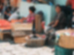 Women in colorful attire sit near wooden crates of tomatoes in a market. A turkey is perched on a basket. Cobblestone ground visible.