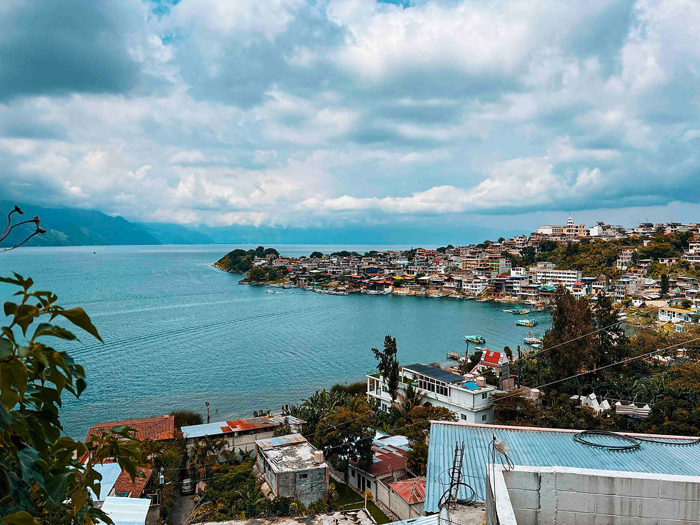Coastal town with colorful buildings on a hill beside a blue lake. Cloudy sky and mountains in the background. Lush vegetation visible.
