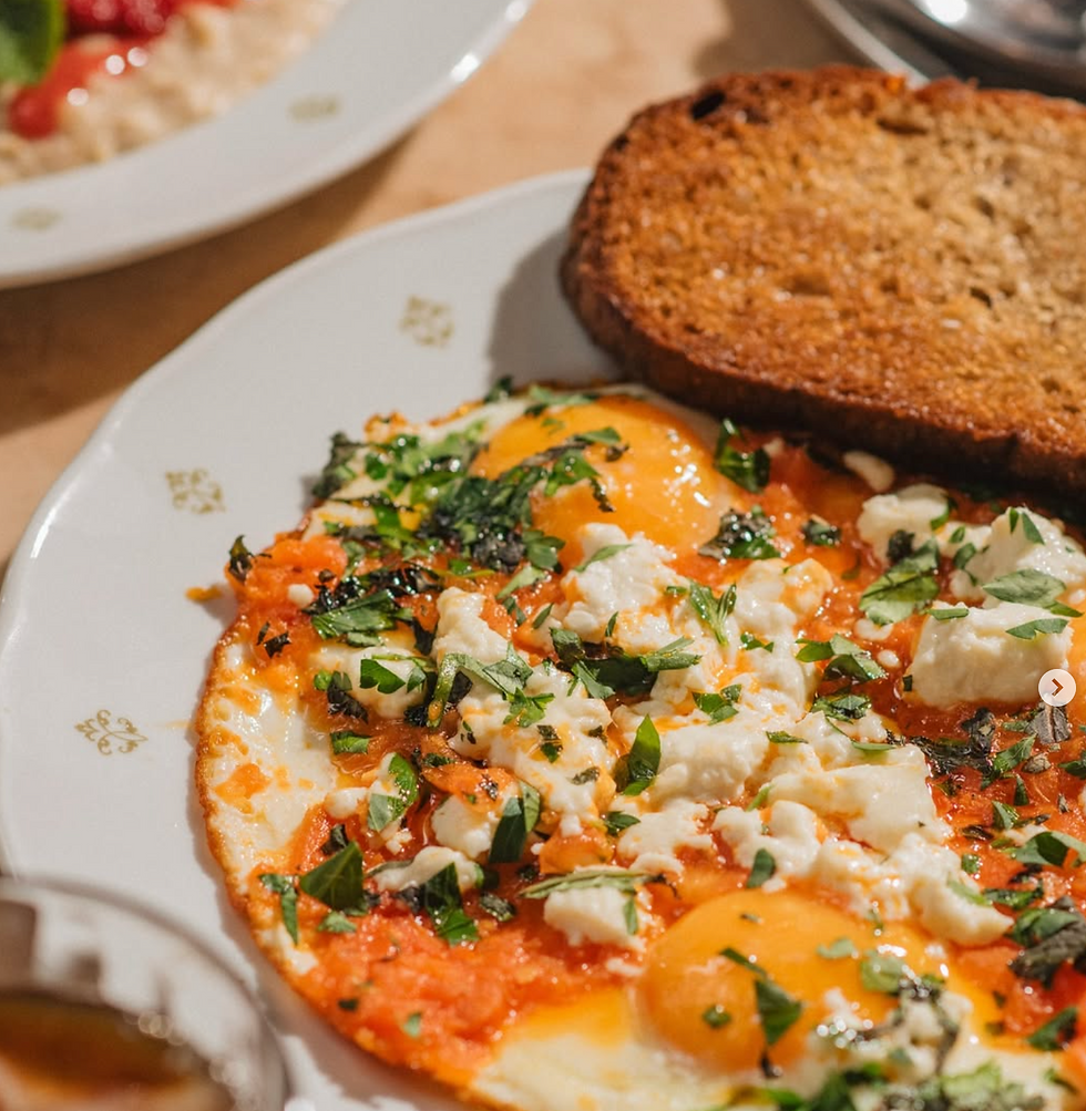 Plate of sunny-side eggs topped with herbs and feta, next to two slices of toast. A bowl of oatmeal with fruit is partially visible. Warm, inviting meal.