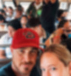 Man in a red cap and woman in a crowded chicken bus. Passengers seated and standing, some looking tired. Close setting with a casual mood.