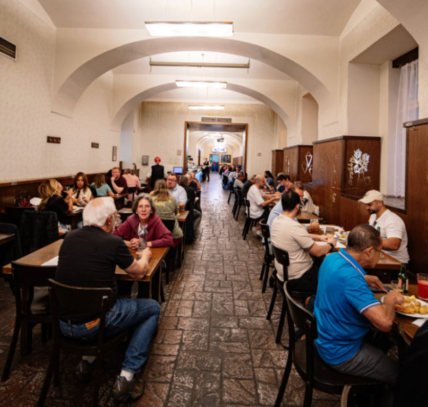 People dining in a bustling, arched-ceiling restaurant. Wooden tables, muted tones, and engaged conversations create a lively atmosphere.