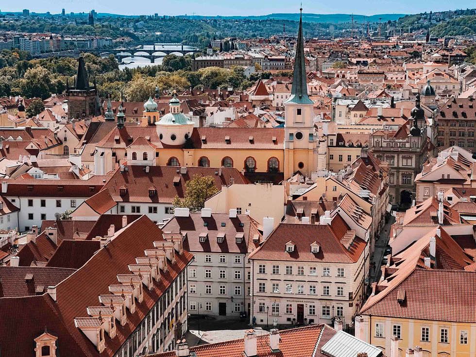 Aerial view of Prague's Lesser Town cityscape with red-roofed buildings, a tall church spire, and distant bridges over a river. Lush greenery surrounds the area.