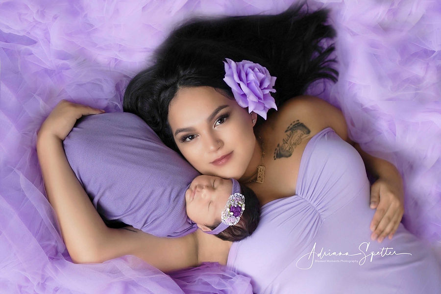 A mother laying down with her newborn baby. The mother is wearing a lilaic gown and surrounded with Lilaic tulle on the floor. Her newborn baby girl is wrapped up in a liliac colored fabric and wearing a headband. The baby is snuggled up on her mother's chin and mama looks up at the camera.