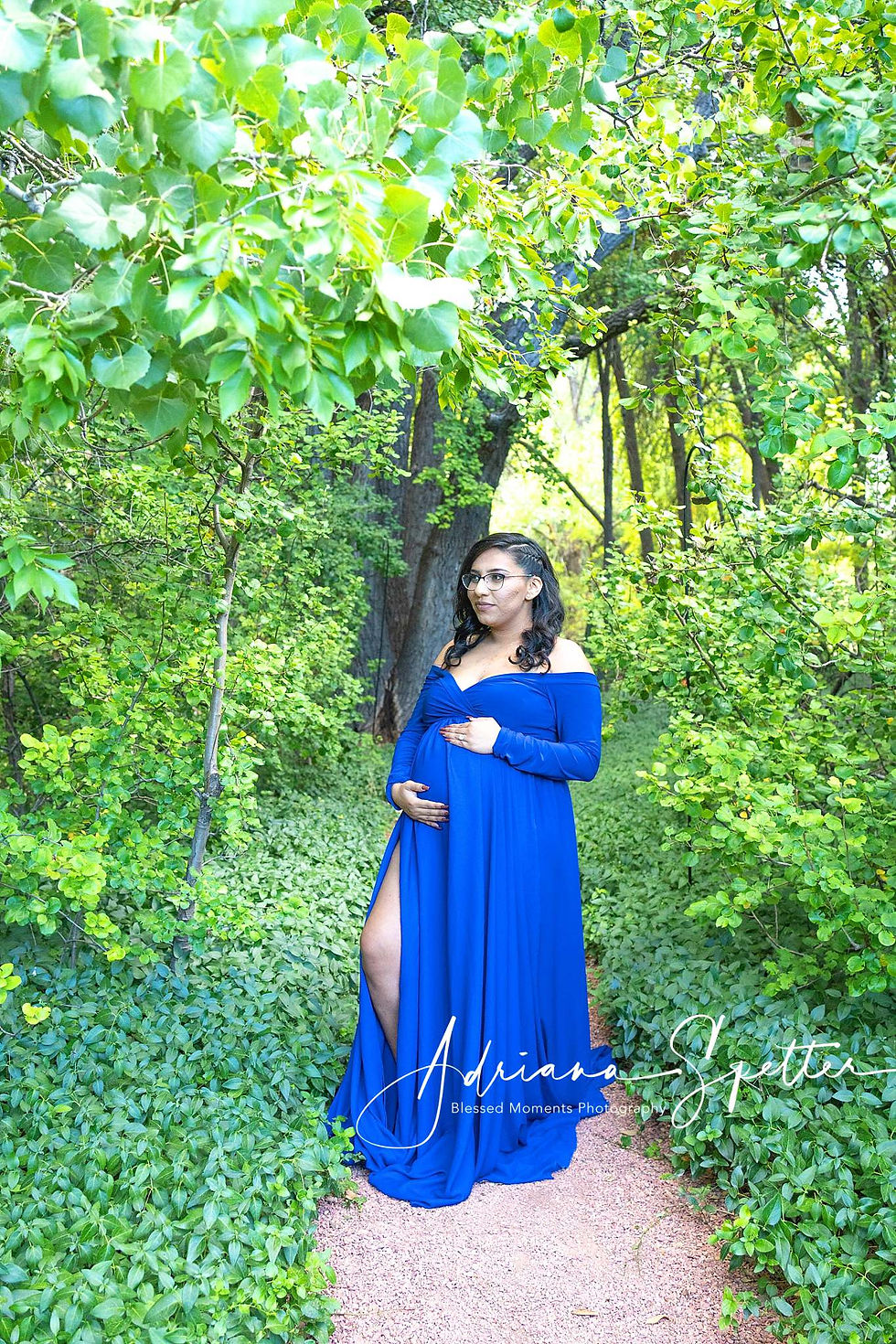 Photo of a young pregnant woman holding her belly while wearing a royal blue maternity gown standing on a gravel path surrounded by trees and periwinkle.