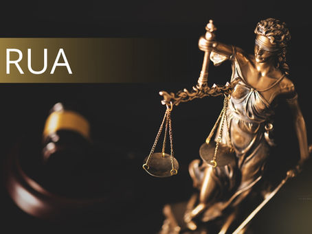 Estatua de bronce de la Dama de la Justicia con una balanza y una espada, con un mazo de juez desenfocado al fondo y el texto “NRUA” destacado a la izquierda sobre un fondo dorado.